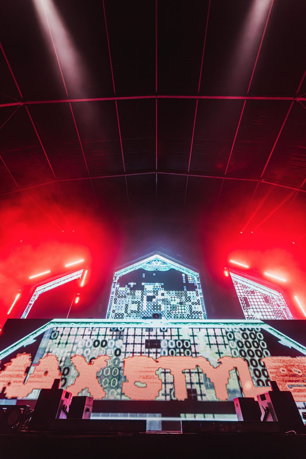 Electronic music concert stage with red lighting and digital graphics displaying across multiple LED screens in panoramic view.