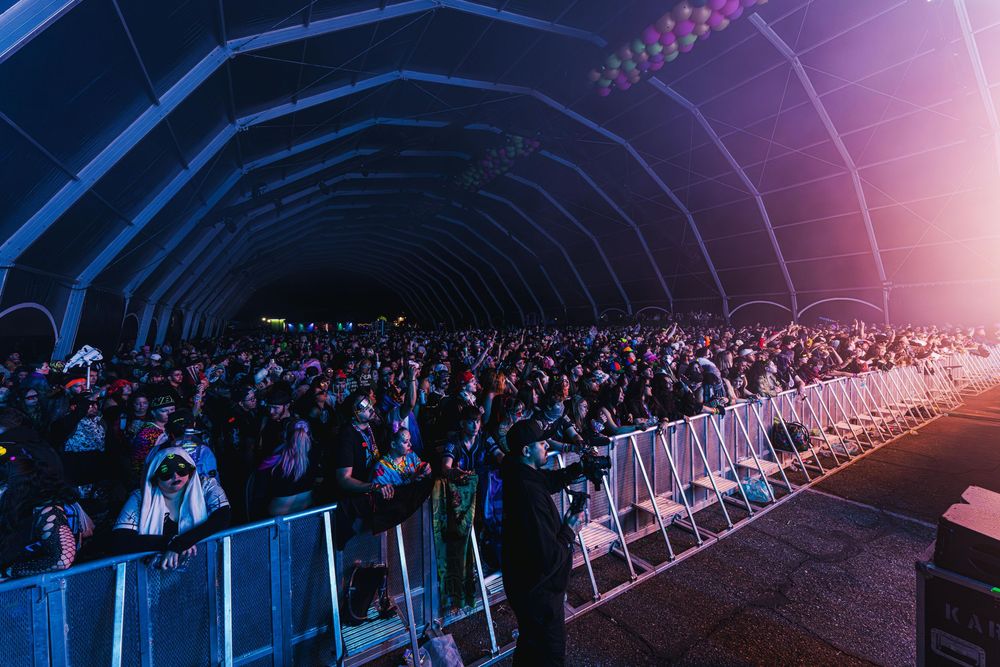 Concert audience sits in rows of white chairs under blue lighting during a large indoor performance event.