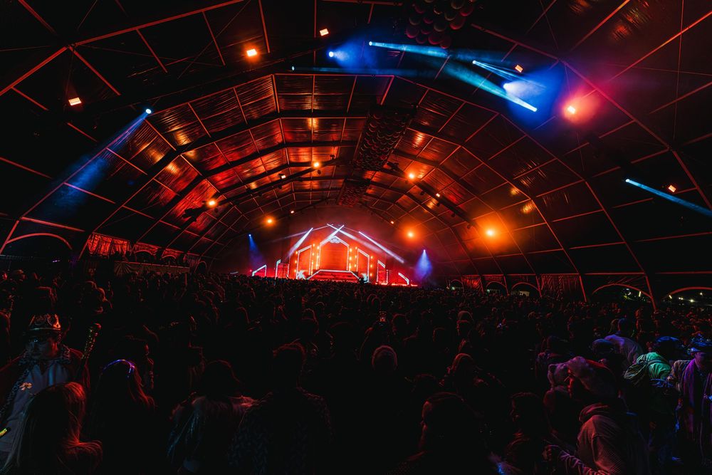 A dark concert venue with illuminated orange and blue lights creating an arch-like pattern above a crowded dance floor.