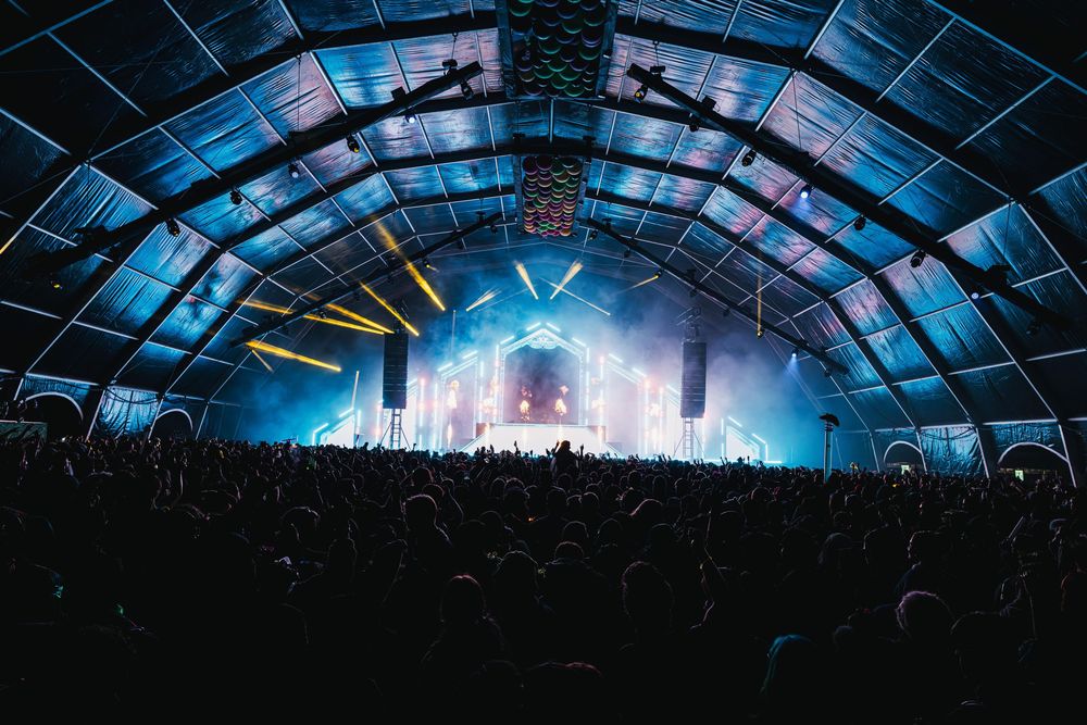 Concert crowd silhouettes under illuminated blue dome ceiling at indoor arena venue during live performance.