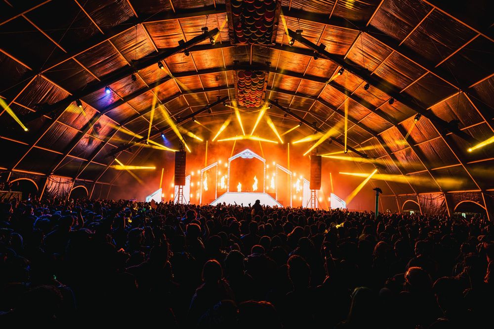 Concert crowd silhouettes beneath colorful stage lighting at an indoor music festival venue.