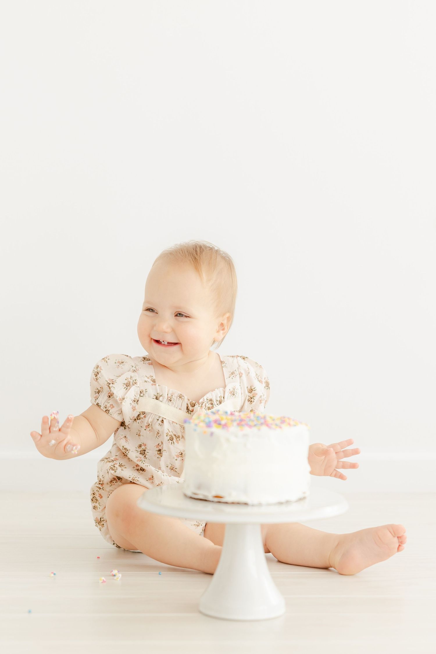 Baby in floral outfit smiling during cake smash photo session with white cake stand | CT studio