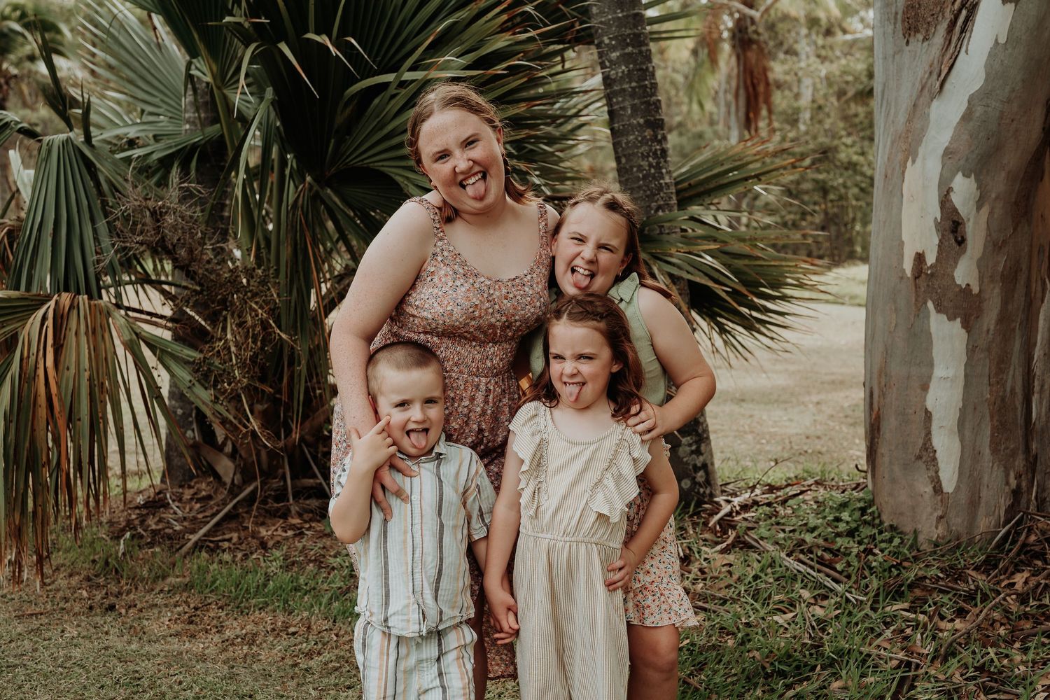 A group of people in coordinating beige and white outfits pose together outdoors among palm trees and greenery.