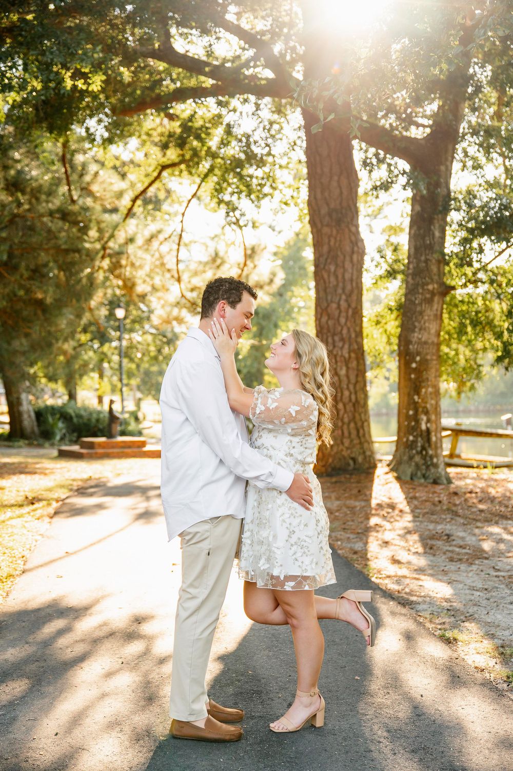 A romantic couple embraces on a sunlit path beneath tall trees during a golden hour photoshoot.