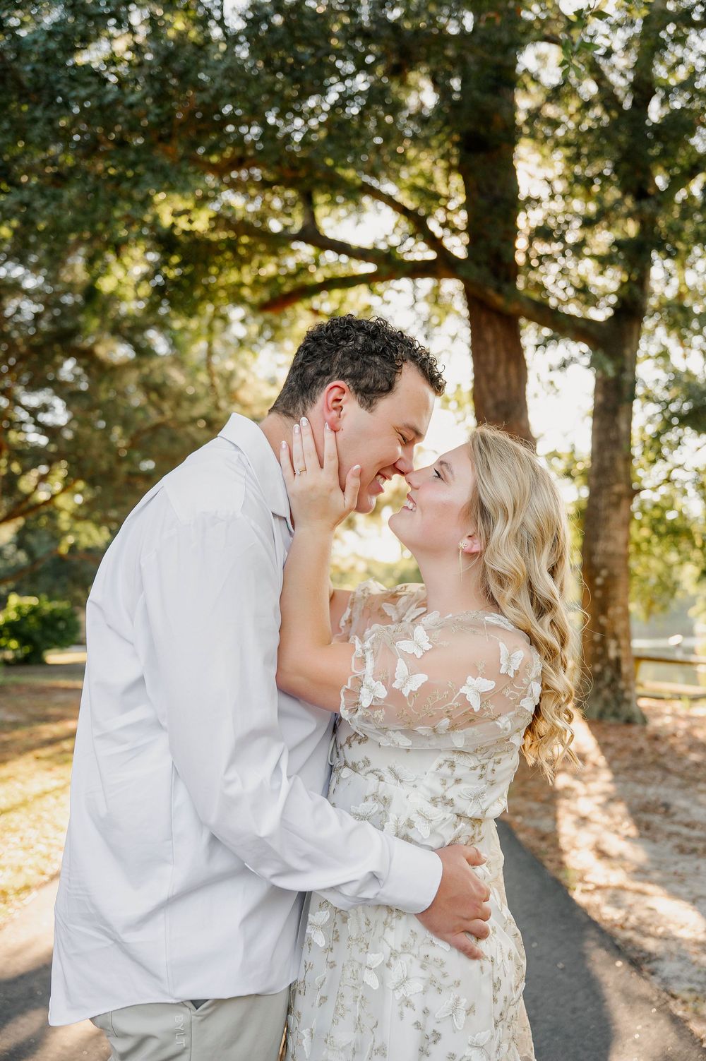 A tender moment between a couple in formal attire beneath sun-dappled trees.