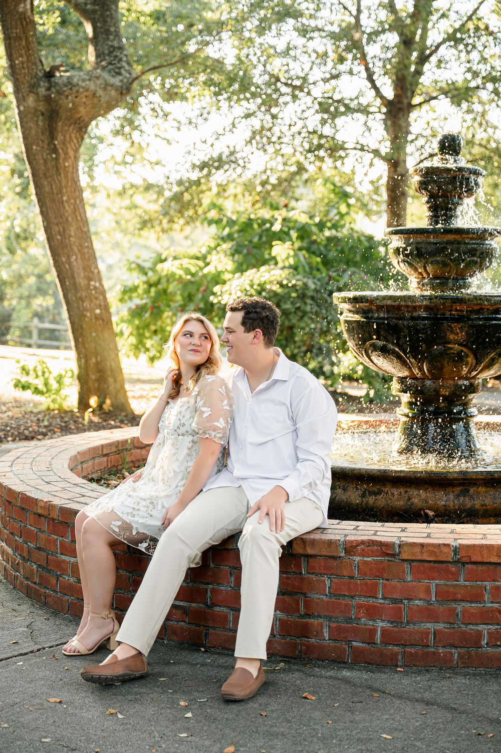 A couple sits together on a brick wall beside an ornate fountain in a garden setting.