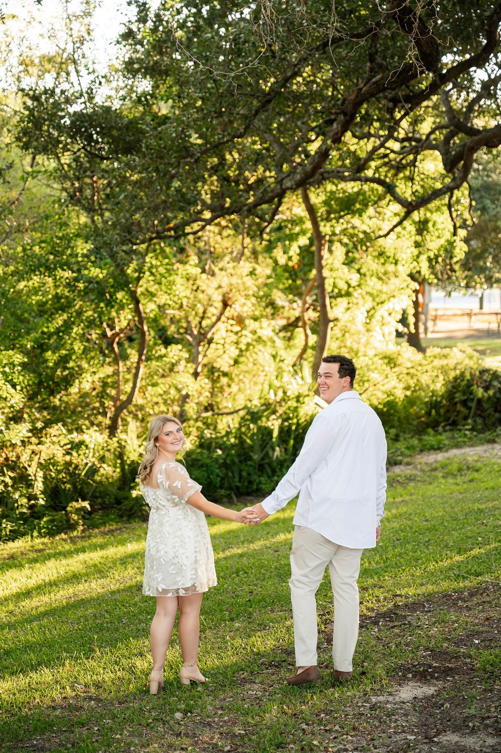 A couple holds hands while dancing in a sunlit garden with trees in the background.