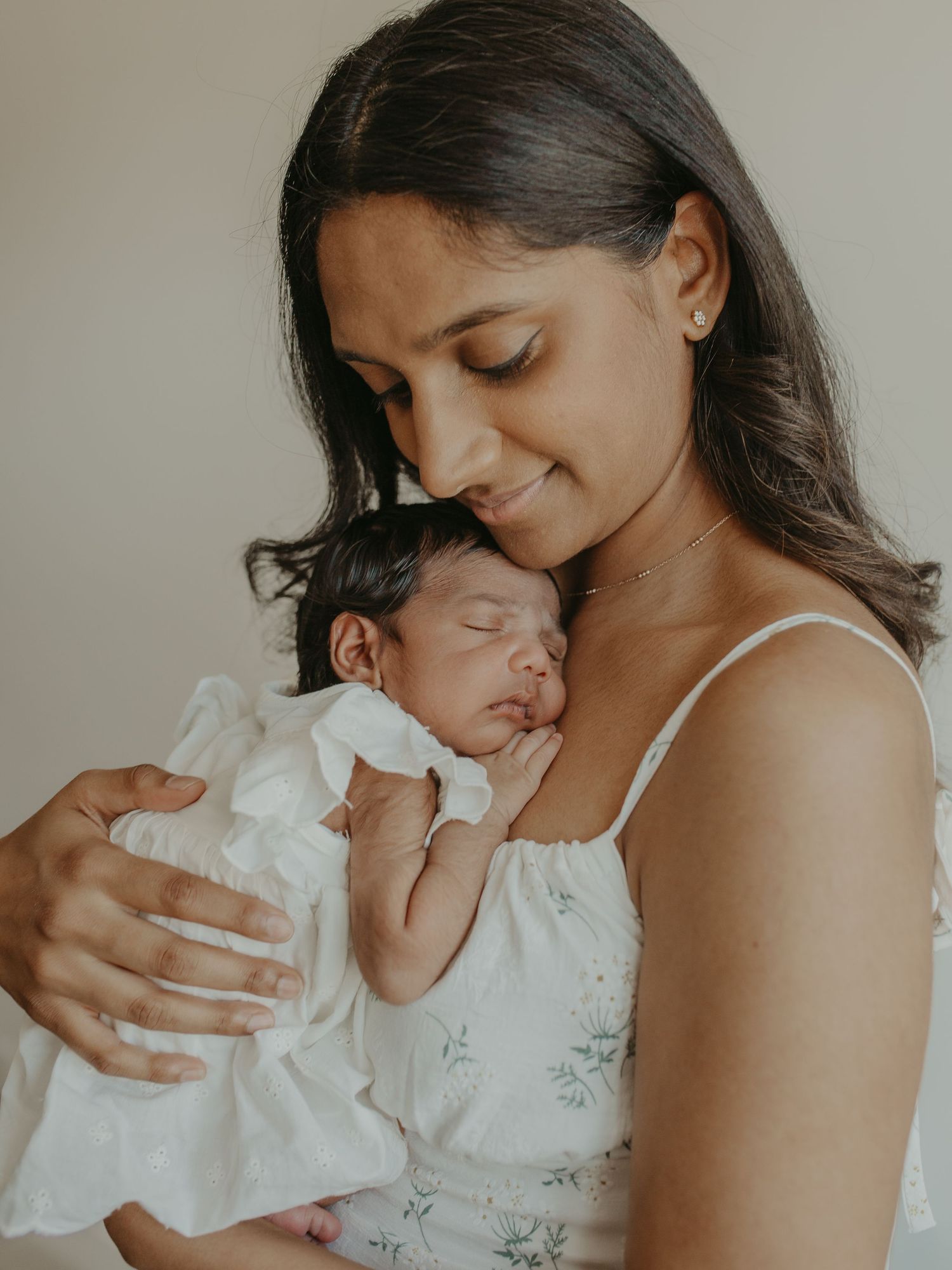 A young mother tenderly holds her sleeping newborn baby dressed in white against her chest.