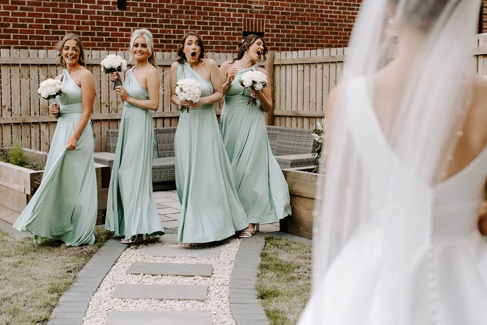 Bridesmaids in matching sage green floor-length dresses walk along a brick pathway holding white bouquets.