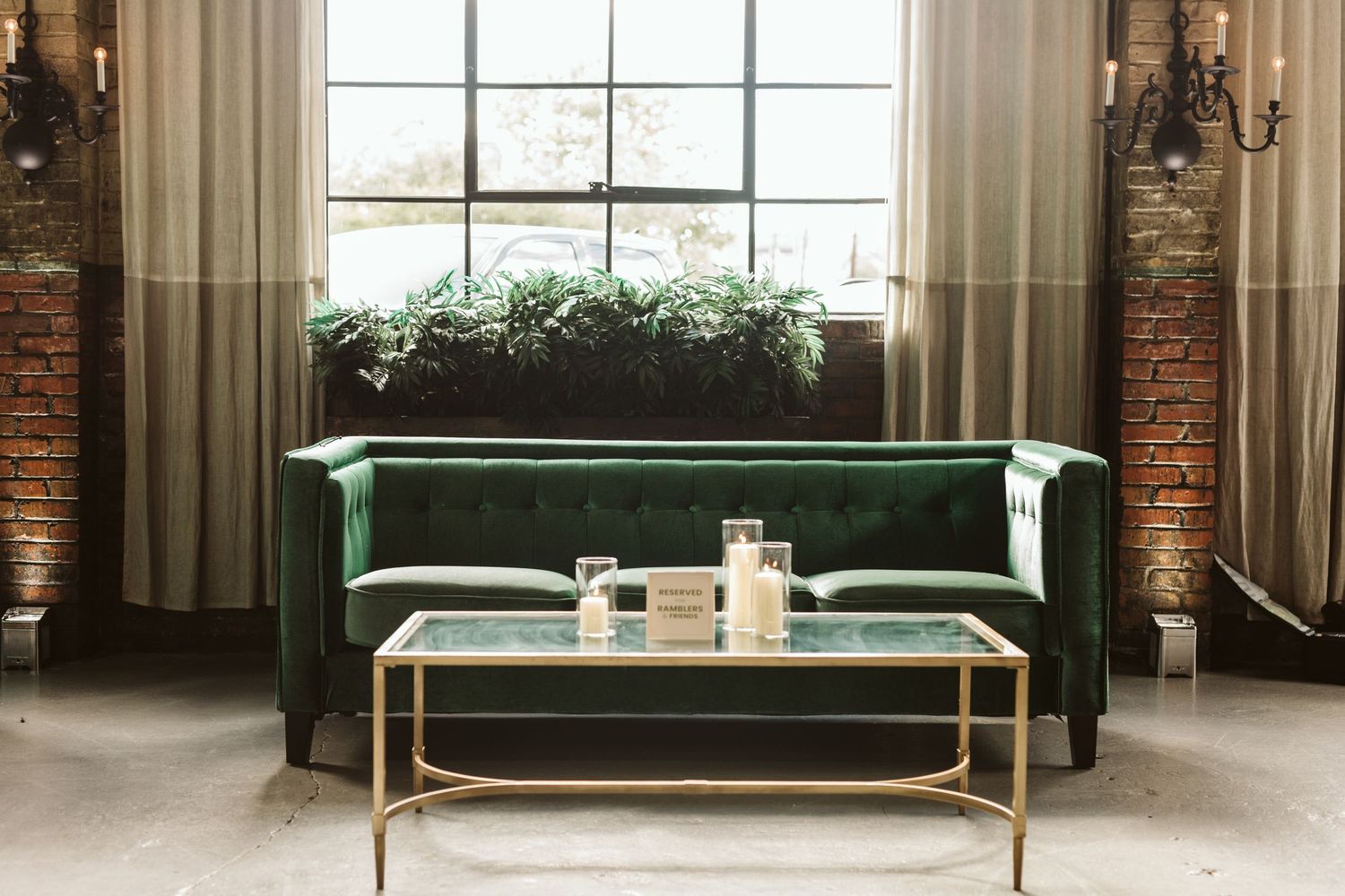 Elegant green velvet sofa with gold coffee table in front of tall windows with natural light and brick accents.