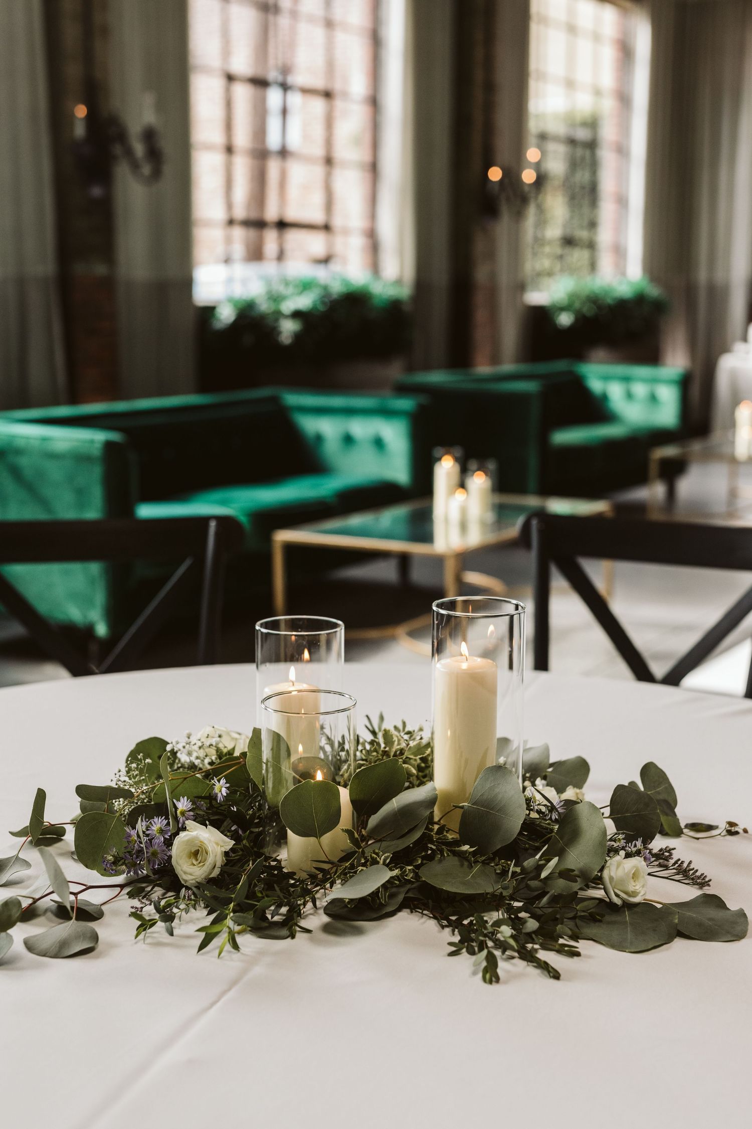 Elegant table centerpiece with white candles and greenery against emerald velvet seating.