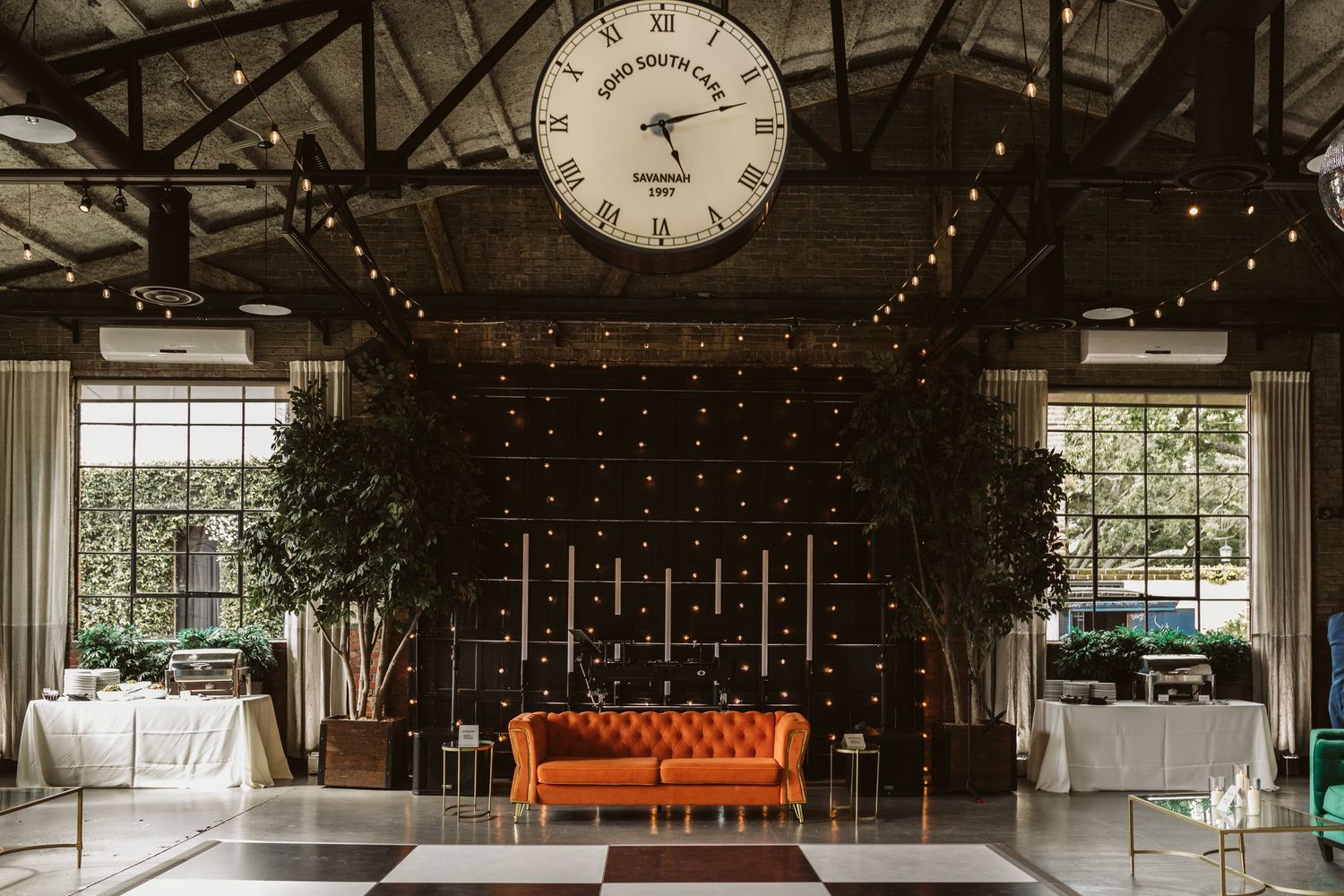 Modern industrial wedding venue with orange couch, large clock, and string lights creating a cozy atmosphere.