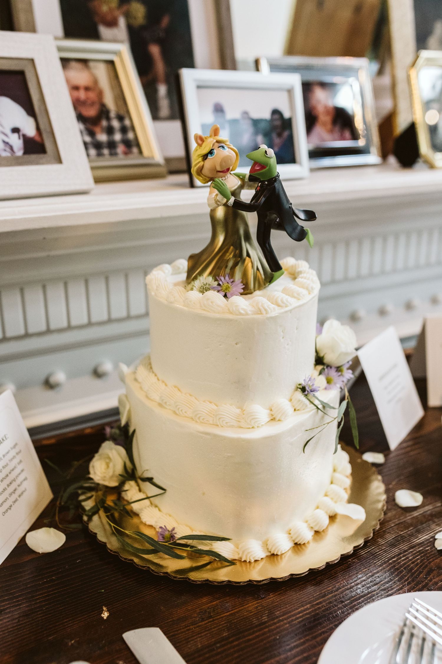 A two-tier white wedding cake topped with Kermit the Frog and Miss Piggy figurines displayed on a wooden table.