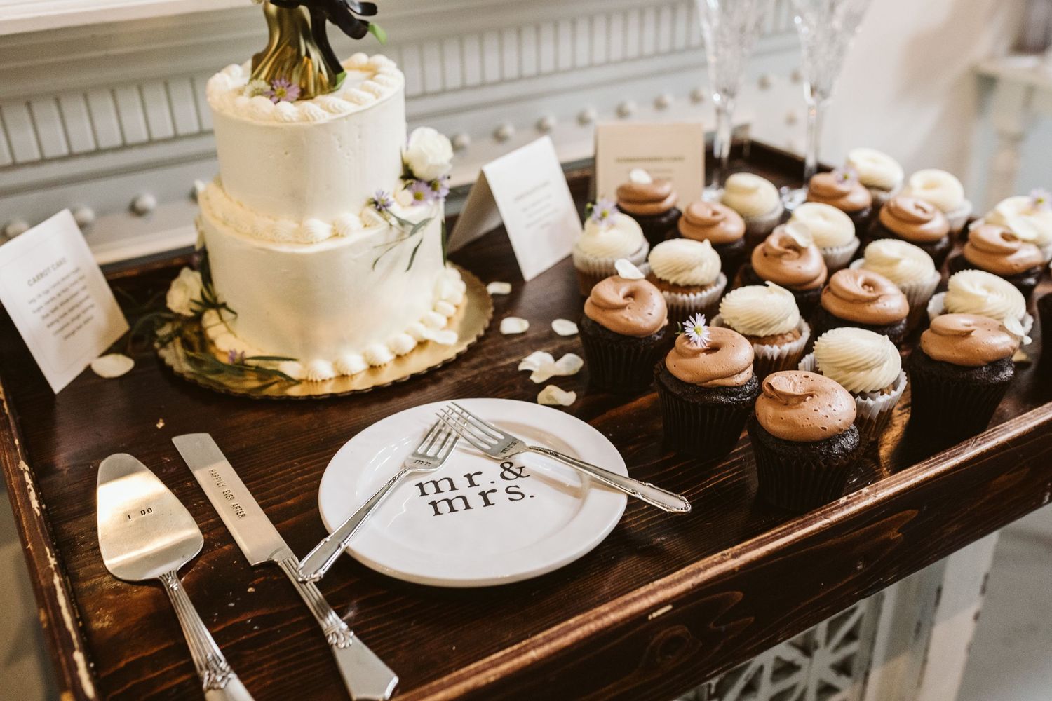 Wedding dessert table with white tiered cake and chocolate cupcakes displayed on dark wood surface.