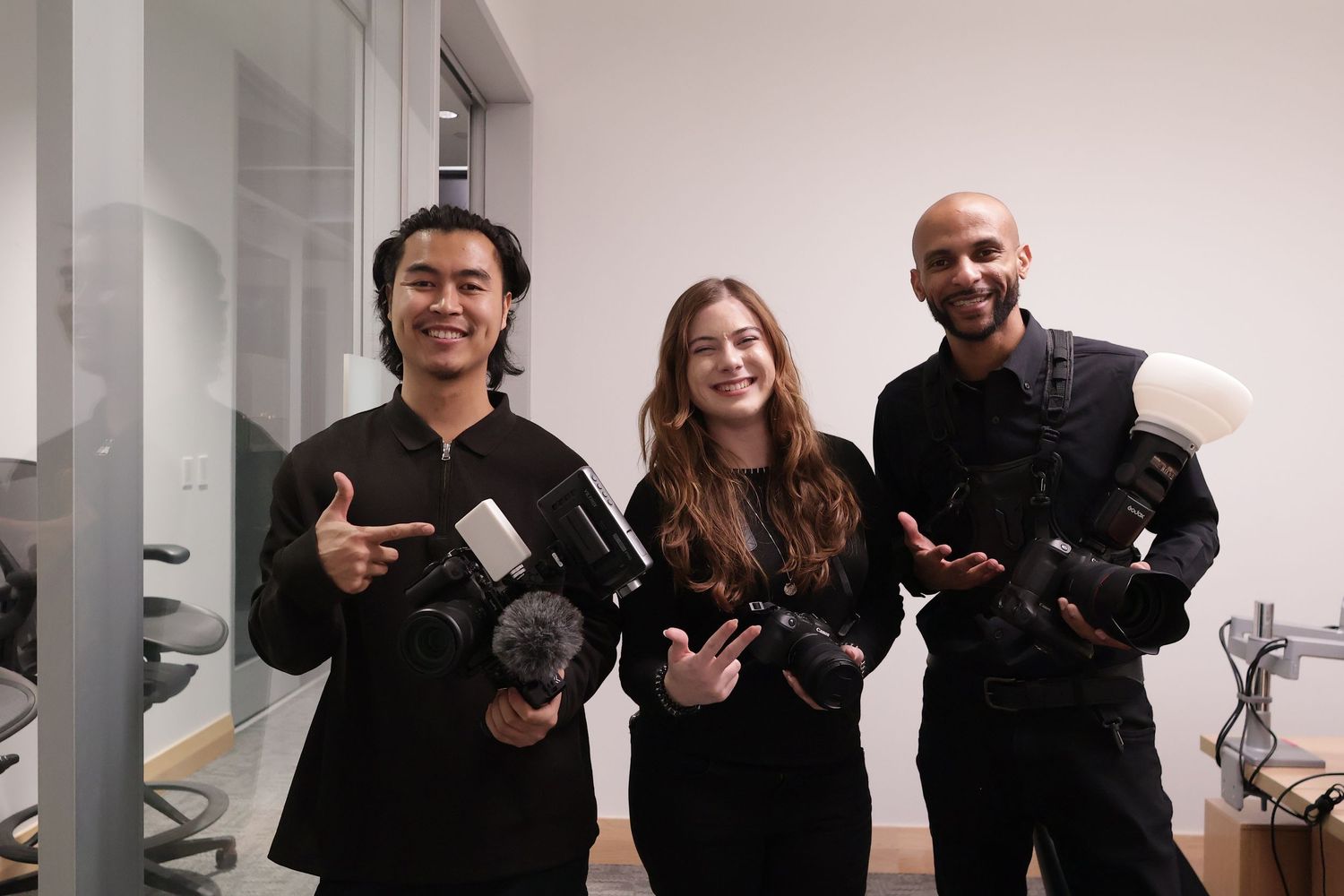 Three people wearing black clothing hold professional cameras and photography equipment in a white studio space.