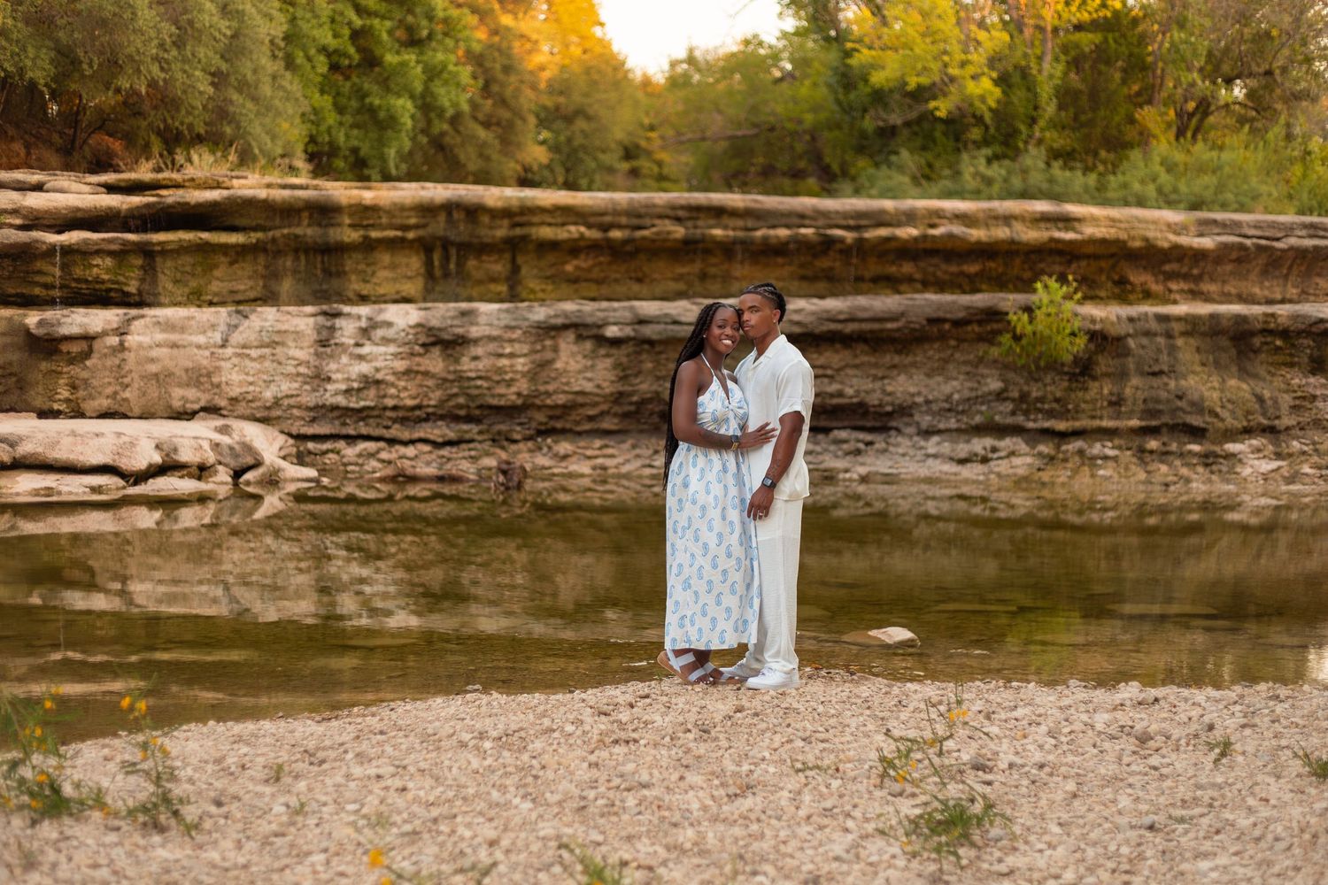A couple in white outfits poses together during their engagement photoshoot along a rocky riverbank at sunset.