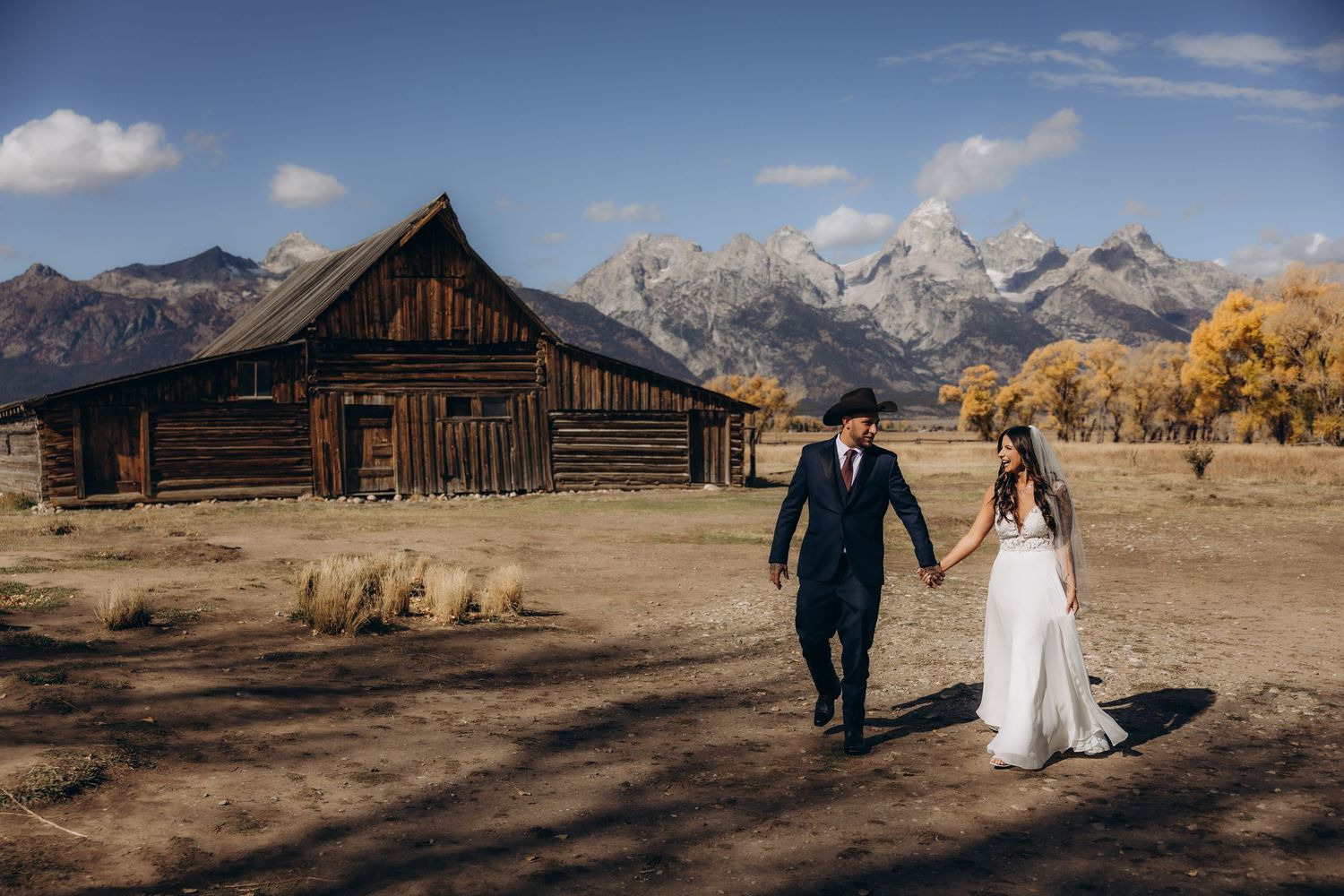 A couple in wedding attire walks through Mormon Row with the Teton mountains rising majestically in the background.