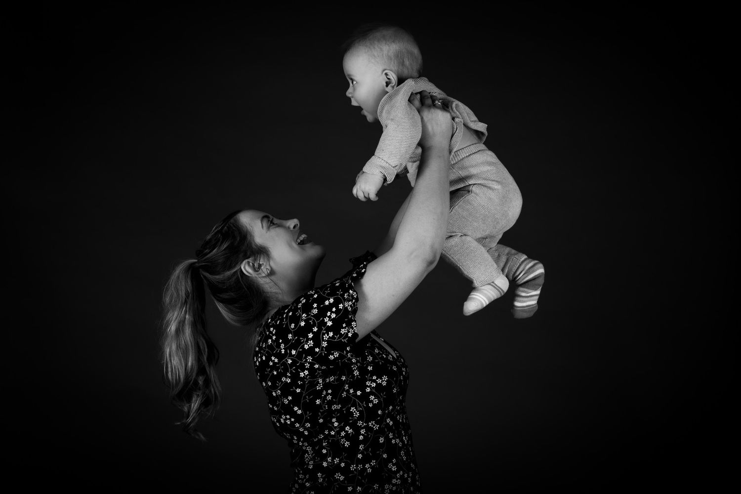 Black and white portrait of a parent lifting a baby in the air against a dark background.