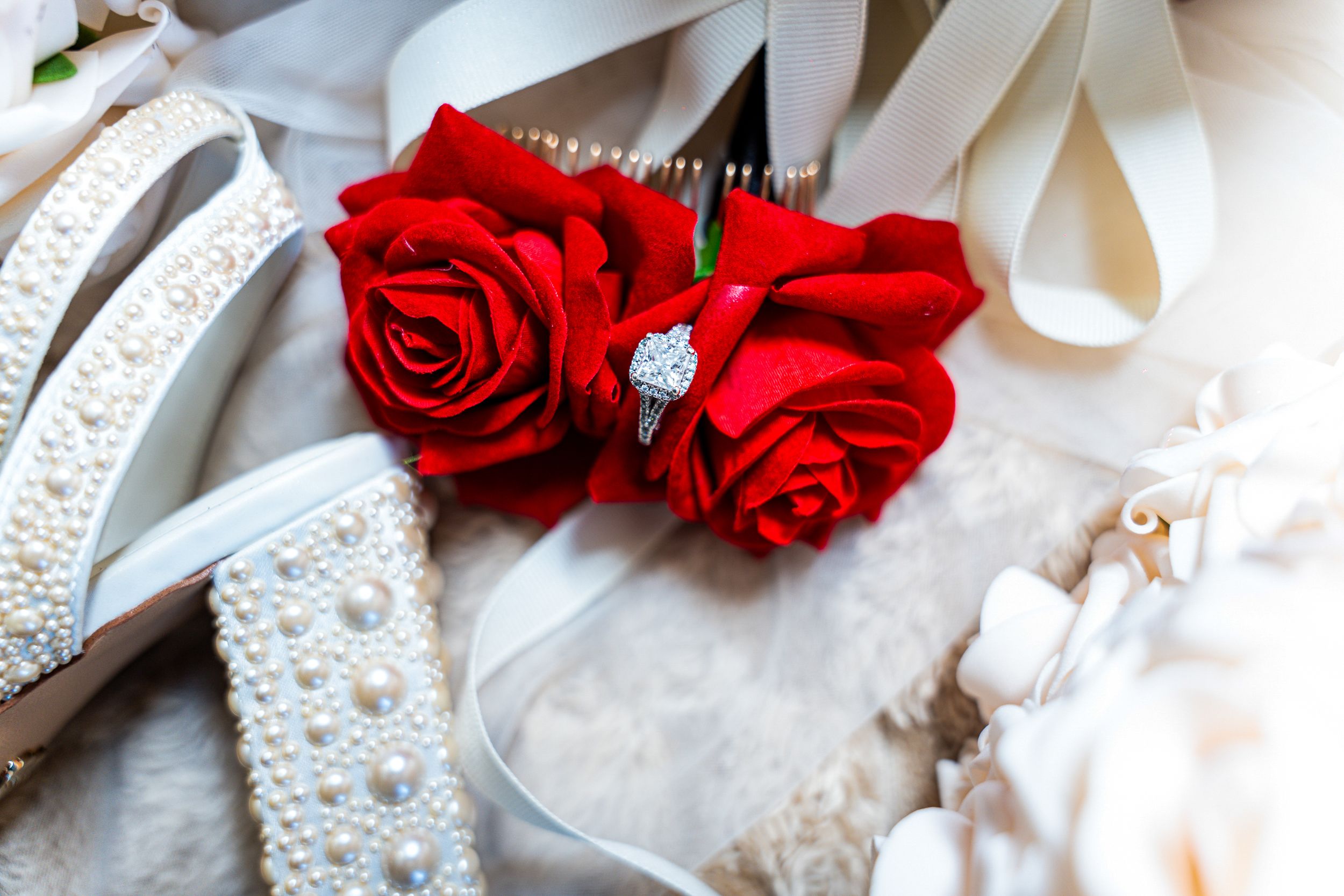 Red roses with a diamond ring nestled between them alongside white lace and pearls in a romantic wedding setting.