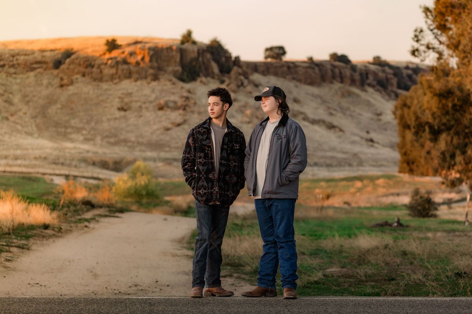 Two people stand together on a hillside path at sunset with warm golden light illuminating the landscape.