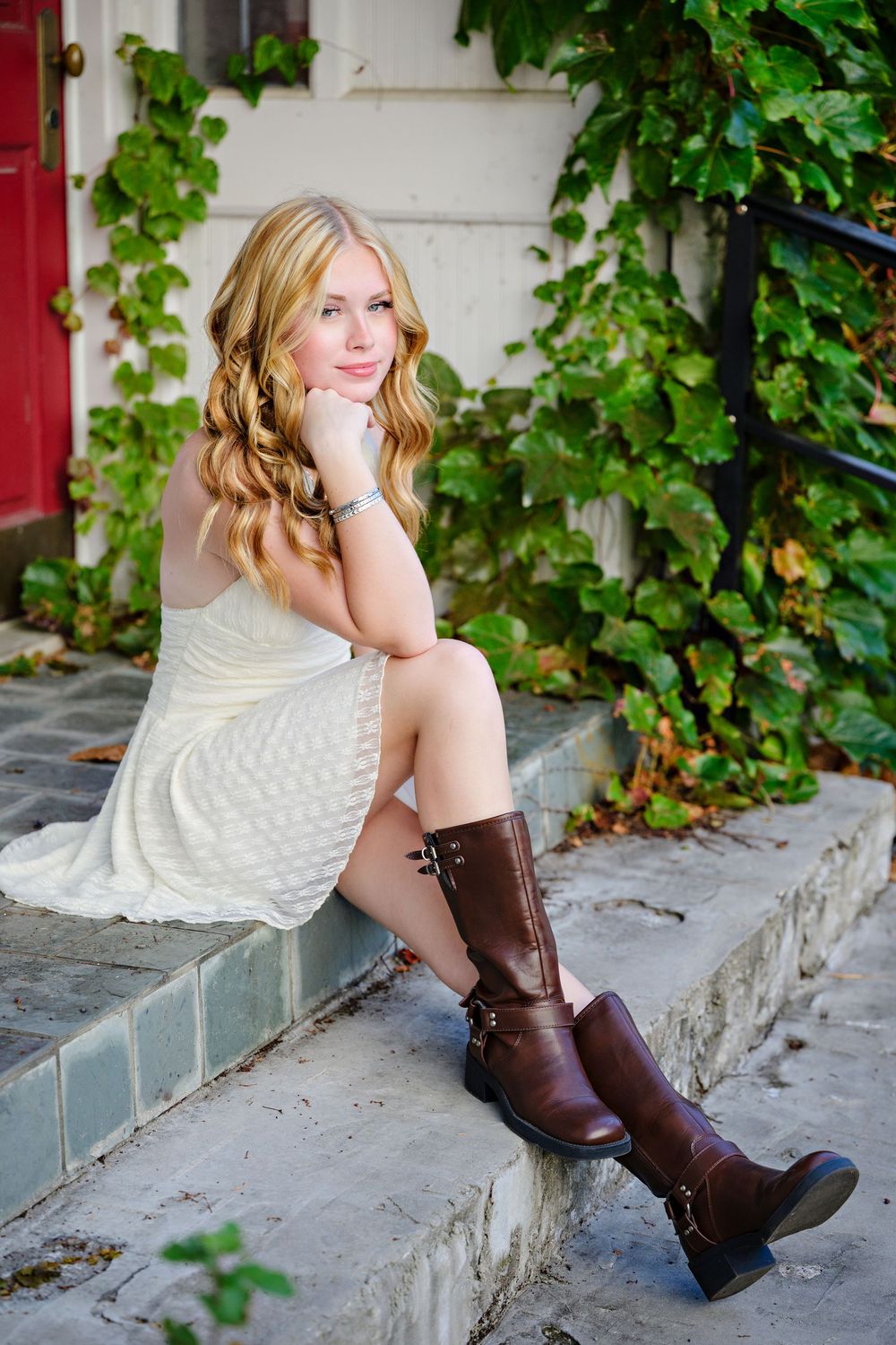 Senior girl in white lace dress and brown leather boots poses on concrete steps with green ivy in background at Columbus Library in Ohio