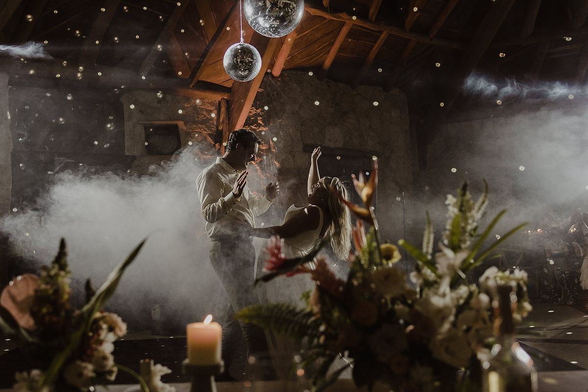 Couple dancing in a misty barn wedding reception with disco ball lights and floral centerpieces.