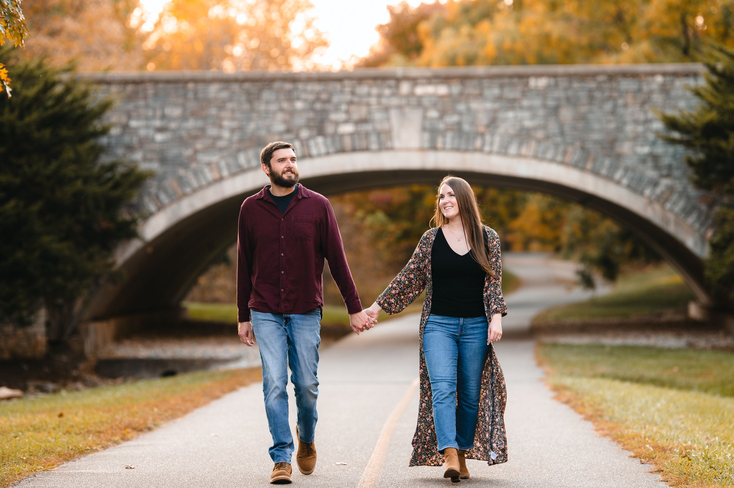 Couple walking hand in hand on a path under a stone bridge during autumn.