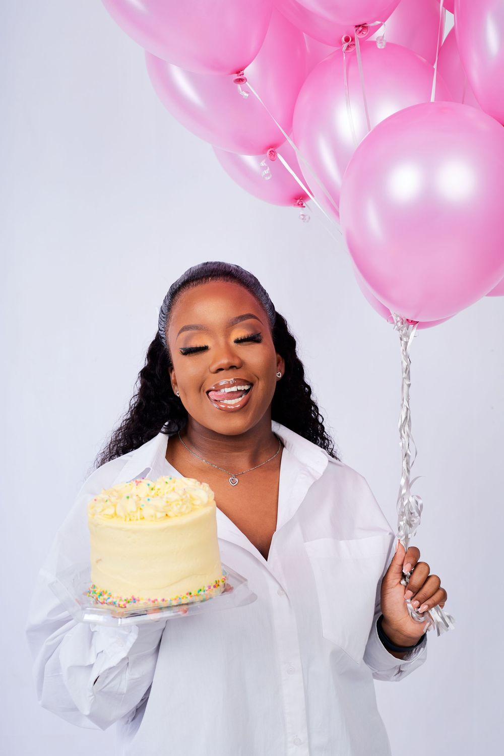 Celebratory photo shoot with pink balloons and a yellow birthday cake against white backdrop.