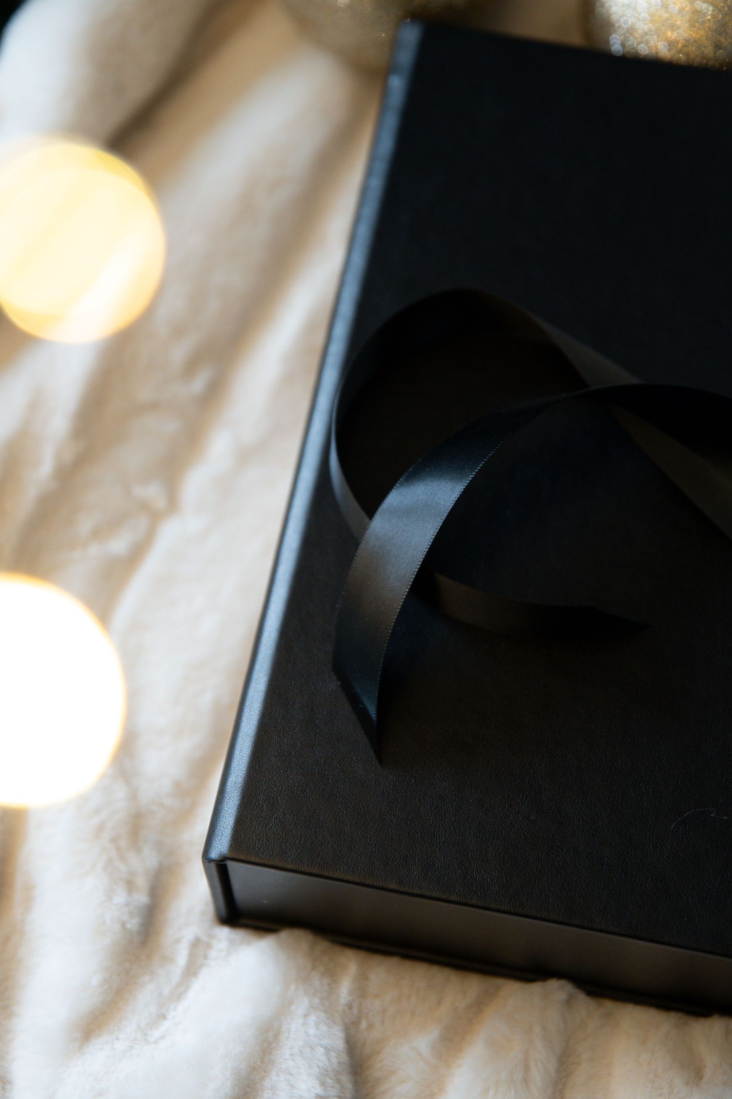 A black gift box with ribbon sits on white fabric, with soft bokeh lights in the background.