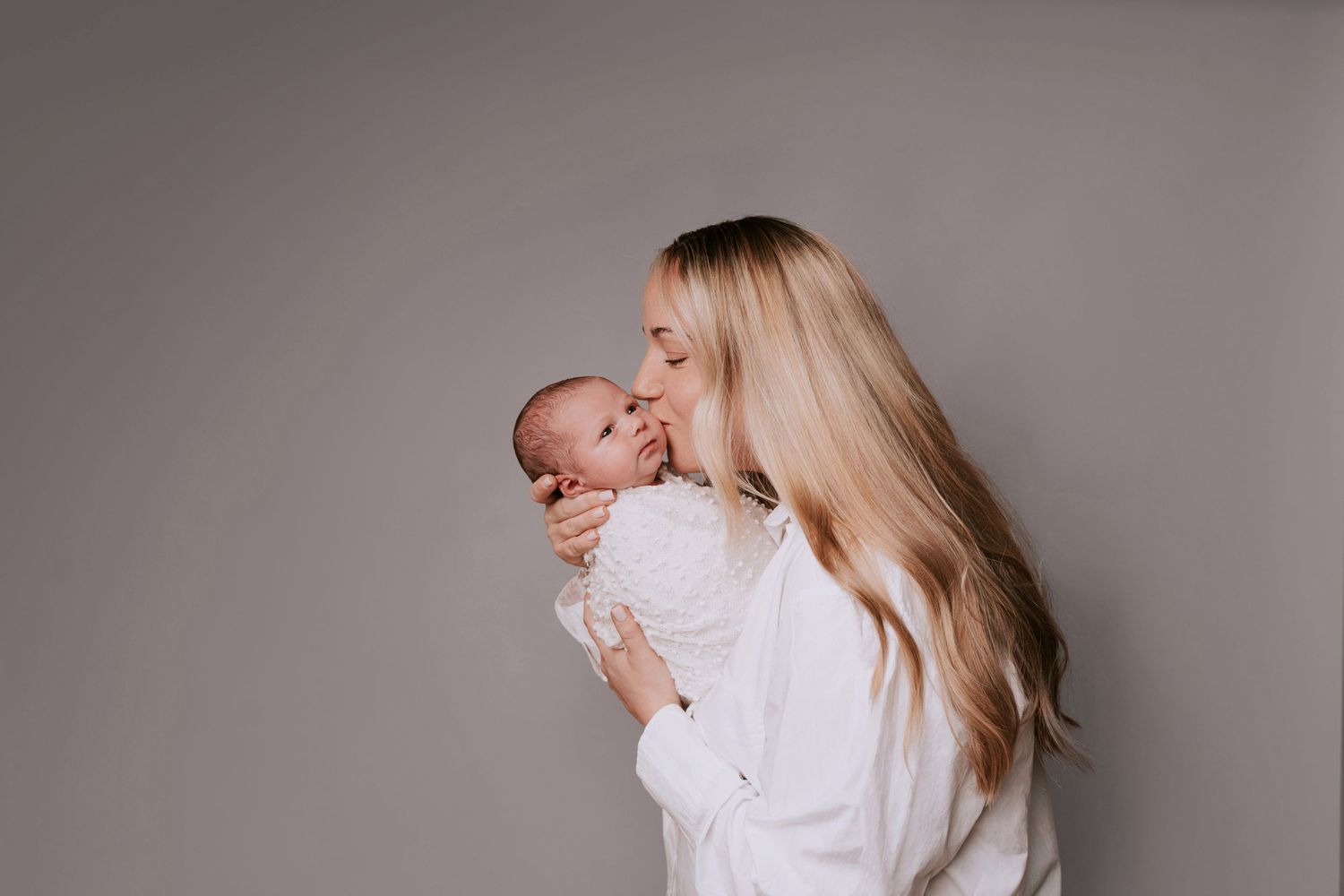 Beautiful mother in a white shirt lovingly kisses the cheek of a smiling baby swaddled in a white blanket, against a grey background.