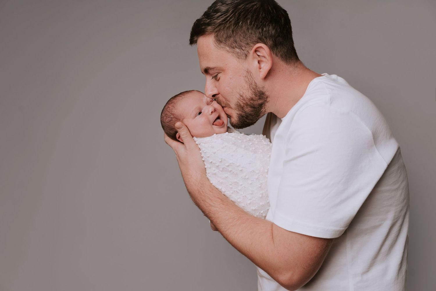 A man in a white shirt gently kisses the forehead of a smiling baby swaddled in a light blanket, against a grey, neutral background.
