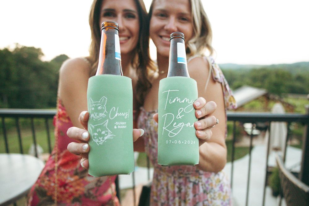 Two friends holding mint green custom wedding drink koozies on an outdoor balcony.