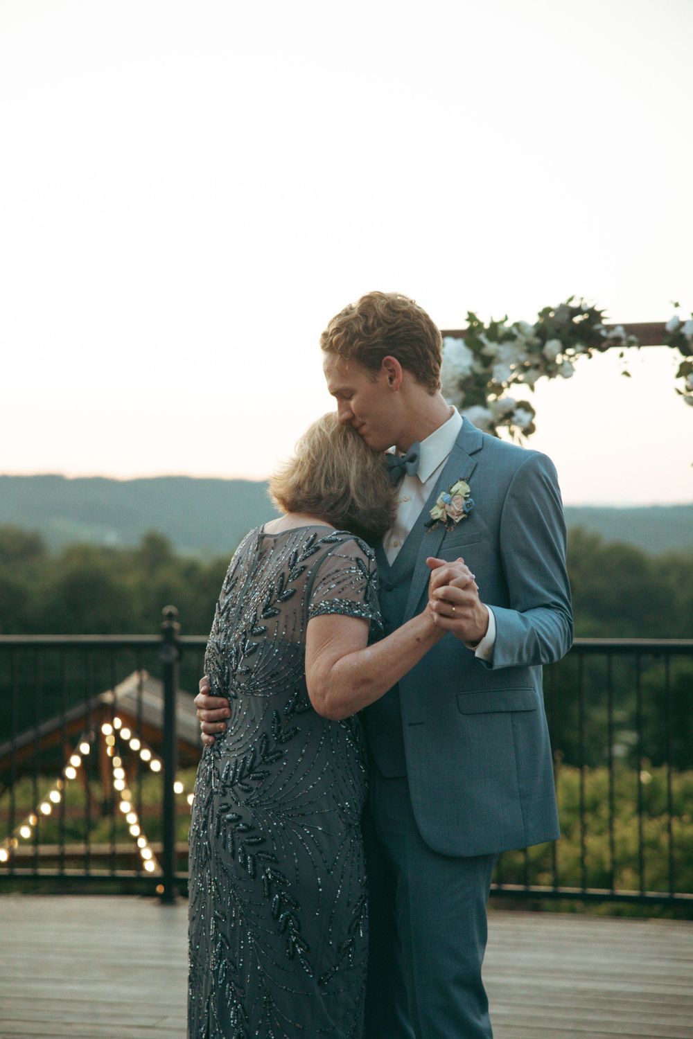 Groom and groom's mother share special dance moment on decorated outdoor terrace at sunset.