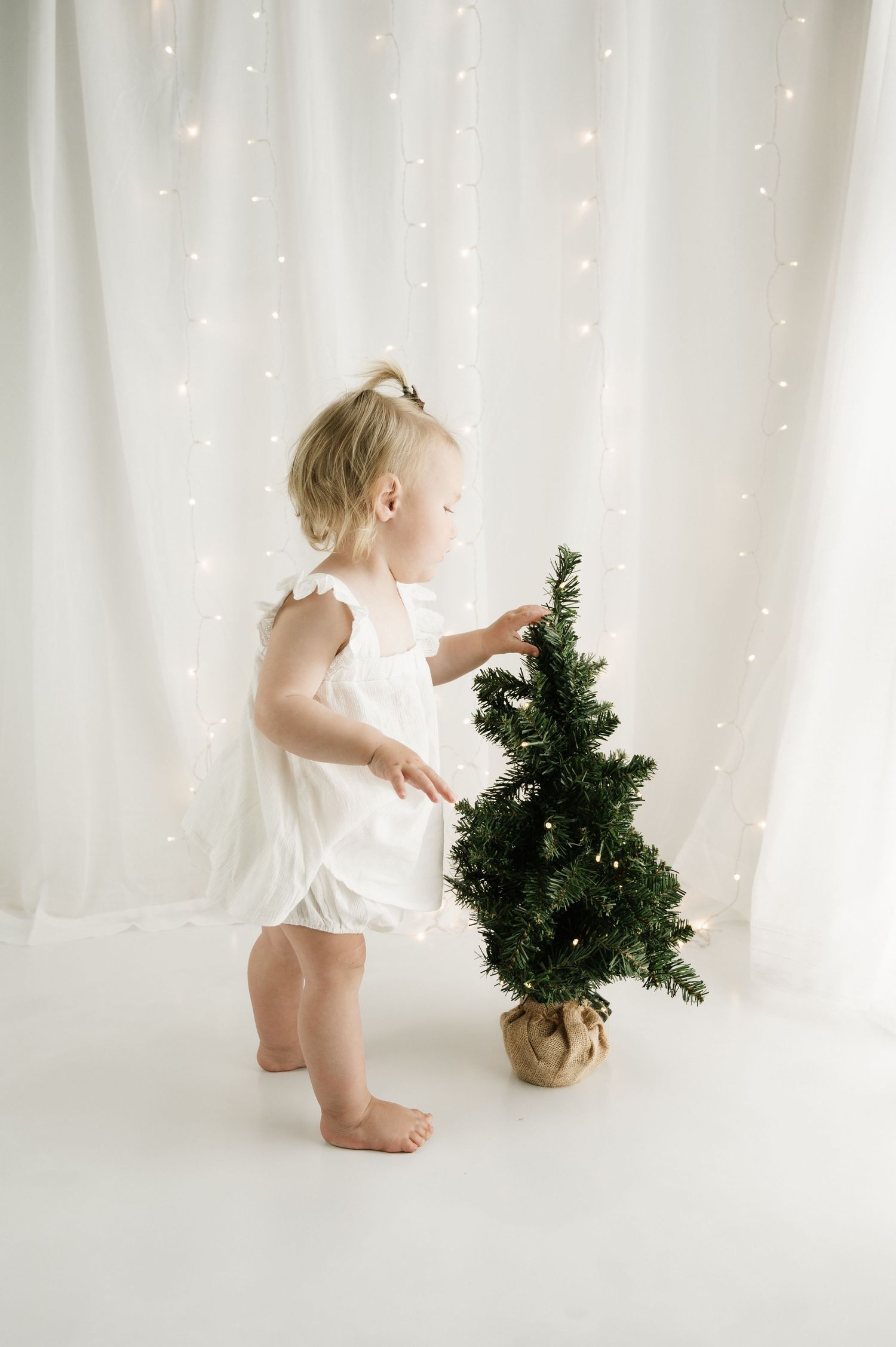 Toddler in white outfit decorating small Christmas tree against sparkly backdrop.