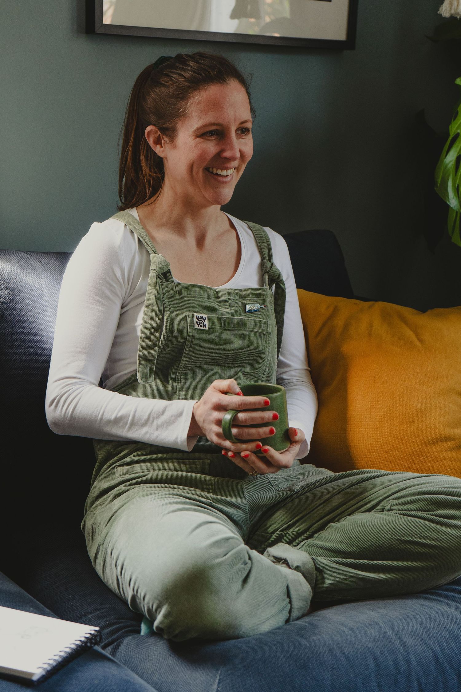 Person in green overalls and white shirt relaxing on yellow couch with mug.