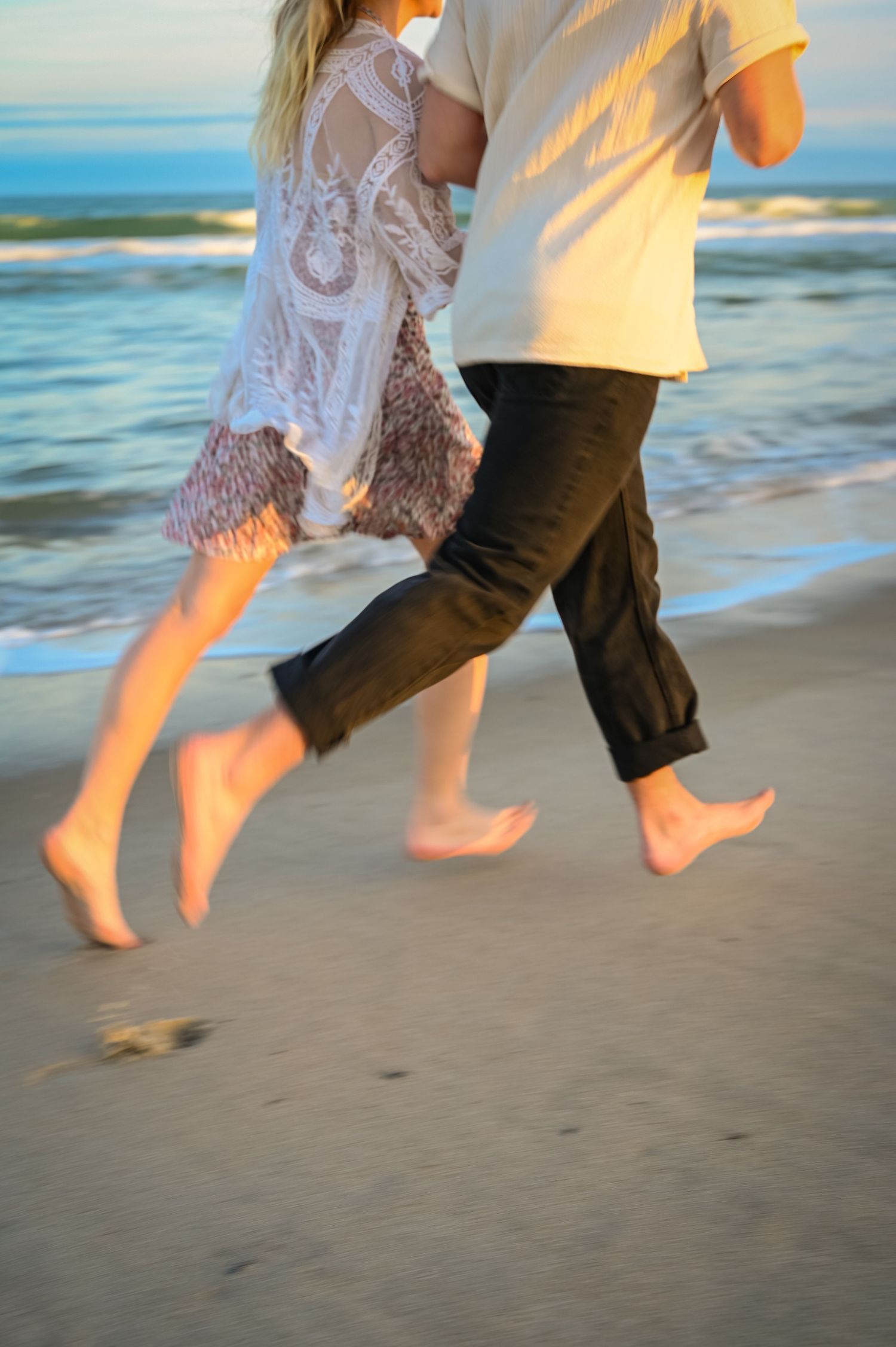 Couple's feet jumping on beach sand with ocean waves in background.
