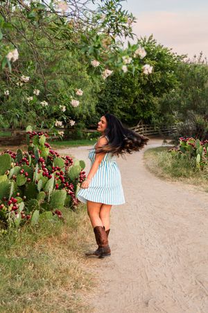 Woman in blue striped dress and cowboy boots twirling on a dirt path near flowering cactus and trees.