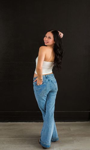 Woman in white crop top and blue flare jeans posing against black wall, looking over shoulder. in studio birthday photoshoot. sweet 16 photo