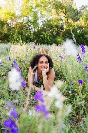 Young woman with curly hair lying in a wildflower field surrounded by purple and white blooms in golden hour light. Senior session North TX