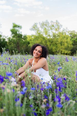 A young woman in a white dress sits smiling in a field of blue wildflowers with green trees in the background. Senior session Richardson TX