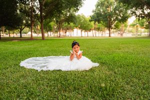 Young girl in white ball gown wedding dress poses on lush green grass in a sunlit park surrounded by trees.