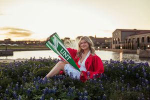 Woman in red jacket holding UNT pennant sits among bluebonnets at sunset near a pond. Senior session Entrada of Westlake