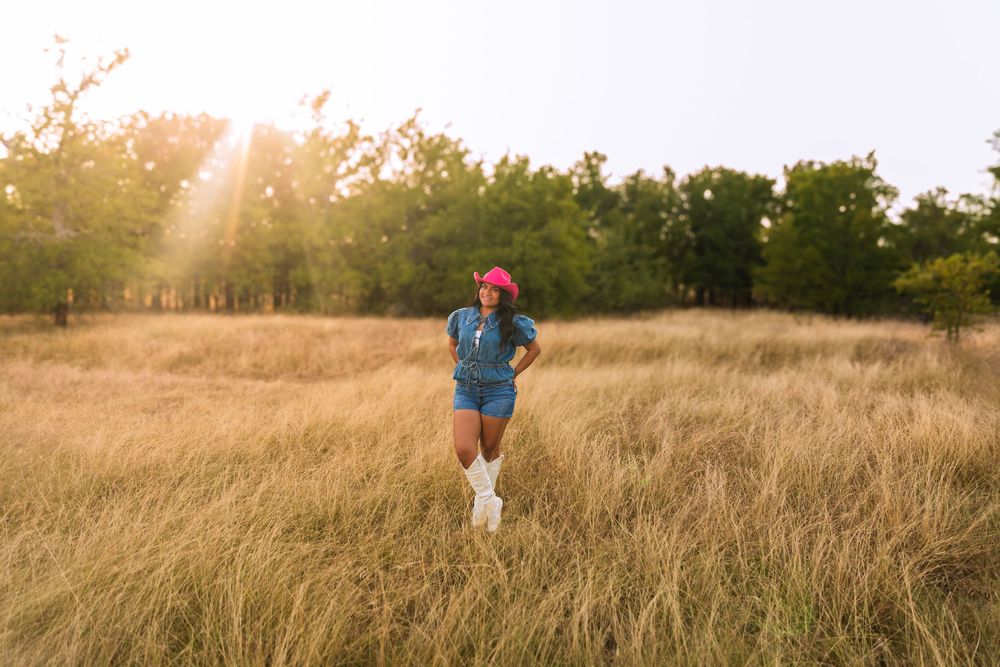 Woman in denim outfit, pink hat, and white boots standing in golden grass field with sunlight streaming through trees.