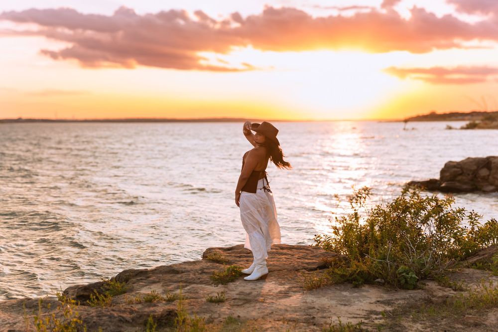 Woman in white pants and hat stands on rocky shore at golden sunset over calm ocean waters.