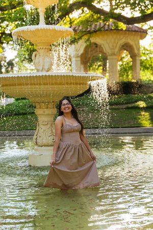 Young woman in elegant maxi dress standing in ornate fountain, smiling, lush garden gazebo background. Senior session Flippen park dallas tx