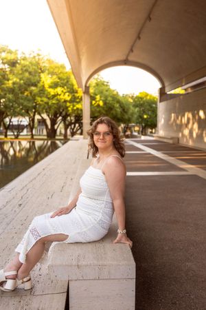 Young woman in white sundress sitting on stone ledge under arched walkway at golden hour. Senior session Kimbell Art Museum Fort Worth Texas