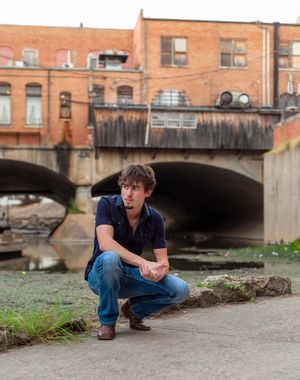Person in a navy shirt and jeans crouches near a brick building with an underpass tunnel in an urban setting. fort worth senior session