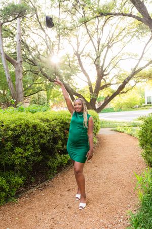 A joyful woman in a green dress tosses her graduation cap on a sunlit garden path. Senior session Flippen Park Dallas Texas