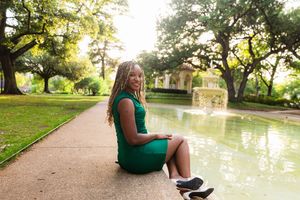 A young woman in a green dress sits gracefully by a fountain in a lush park with sunlit trees. Senior session Dallas Flippen Park Texas