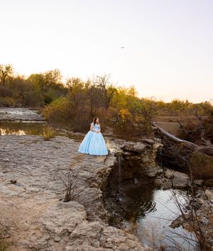 A person in a light blue gown poses on rocky terrain near a water feature during golden hour sunset. Saginaw waterfall trail photos